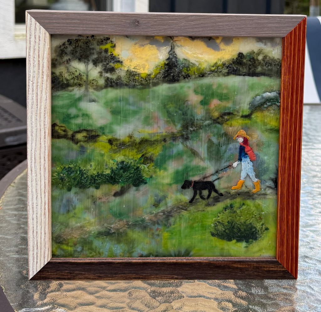 Original encaustic painting of a figure in yellow boots and red jacket walking a black dog through a green rainy landscape, professionally framed in two-tone wood